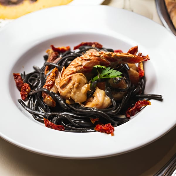 Black squid ink pasta with shrimp, sun-dried tomatoes, and parsley in a white bowl.