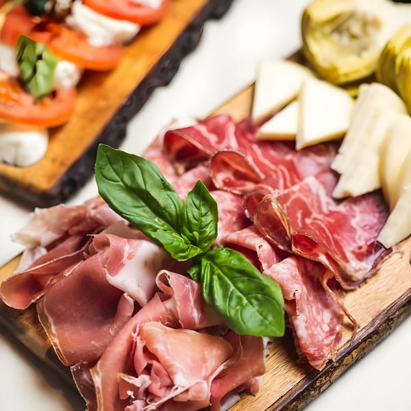 Italian charcuterie board with prosciutto, salami, cheese, basil, and caprese salad in the background.