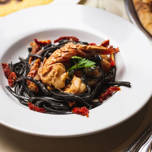Black squid ink pasta with large shrimp, sun-dried tomatoes, and parsley in a white bowl.