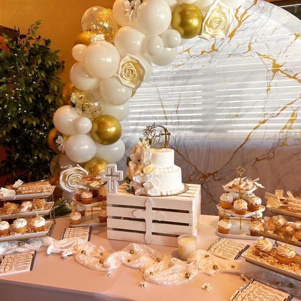 A dessert table with a two-tiered cake, cupcakes, and pastries, decorated with white and gold balloons.