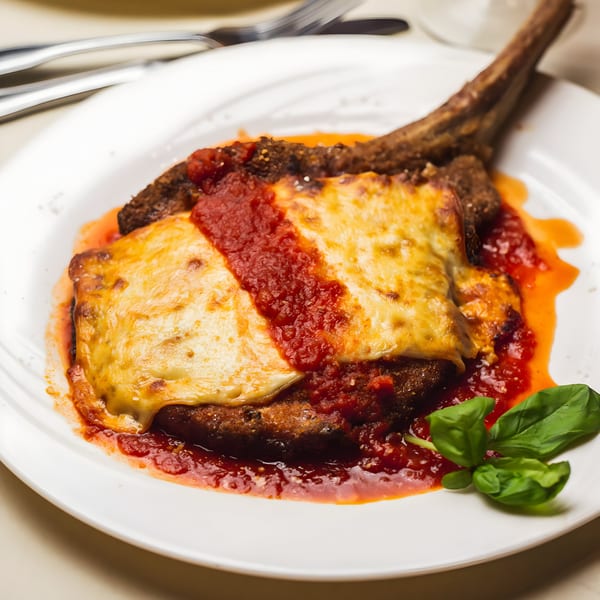 Veal chop parmesan with melted cheese, rich red sauce, and fresh basil on a white plate.
