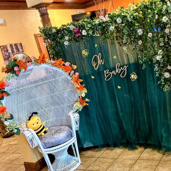 White wicker chair with a bee plushie, adorned with flowers, in front of a green "Oh Baby" backdrop.