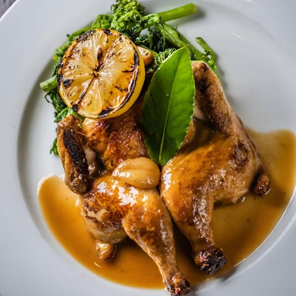 Roasted chicken with broccolini, garlic cloves, a bay leaf, and a grilled lemon on a white plate.