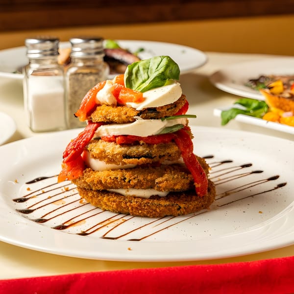 Stacked fried eggplant, mozzarella, roasted red peppers, and basil on a white plate with balsamic glaze.